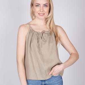 May include: A sleeveless beige top featuring a gathered neckline with a central tie closure. The garment has a relaxed, loose-fitting silhouette and is paired with blue denim jeans. The fabric has a visible, textured weave throughout the piece.
