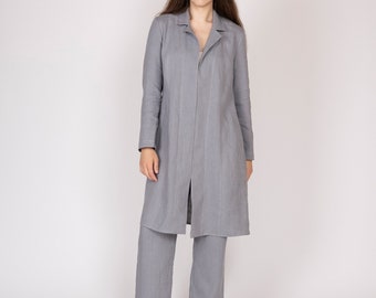 May include: A light gray linen suit, consisting of a long, open-front coat and matching wide-leg pants. The outfit is made of a soft, textured fabric. The model is standing barefoot against a white background.