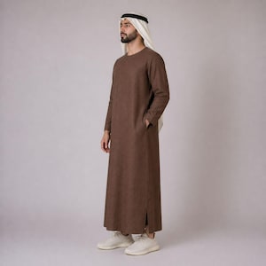 Puede incluir: Un thobe marrón, una prenda árabe tradicional, con mangas largas y bolsillos laterales. El thobe se usa con un tocado blanco y negro y zapatillas blancas. La prenda está hecha de una tela texturizada.