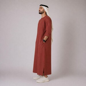 May include: A rust-colored, long-sleeved thobe with side pockets and a split hem. The traditional garment is paired with a white head covering and white sneakers. The thobe is a loose-fitting, ankle-length robe.