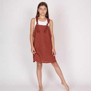May include: A rust-colored linen overall dress with a front pocket and adjustable straps. The dress is worn over a white tank top. The model is standing barefoot against a white background.