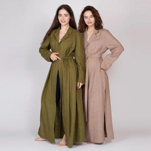 May include: Two women wearing long linen dusters. One duster is olive green, the other is a light brown. Both dusters have a tie belt at the waist and a slit up the front. The dusters are floor length.