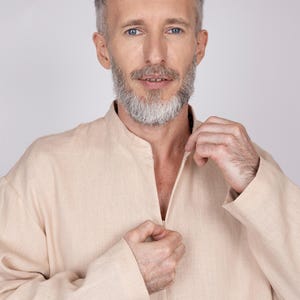 May include: A light beige linen shirt with a mandarin collar and a partial zipper closure. The long-sleeved shirt is being zipped up by the wearer. The shirt is a neutral color and appears to be made of a lightweight fabric.