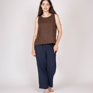 以下が含まれることがあります： 白い背景に立っている、茶色のノースリーブの麻のトップスと濃い青の麻のパンツを着用した女性です。