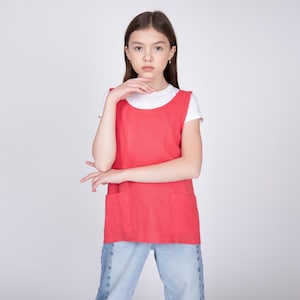 May include: A sleeveless, coral-red apron-style top with two front pockets. The top is worn over a white t-shirt and paired with light blue jeans. The outfit is simple and casual.