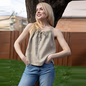 May include: A beige linen tank top with a drawstring neckline and a small bow detail. The top is paired with blue jeans. The model is leaning against a tree.