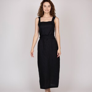 Könnte beinhalten: Schwarzes Leinenkleid mit quadratischem Ausschnitt und breiten Trägern. Das Kleid hat ein figurbetontes Oberteil und einen langen, fließenden Rock. Ein passender Gürtel betont die Taille. Das Model ist barfuß und lächelt.