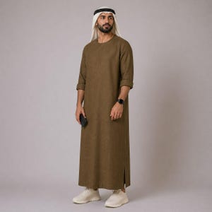 Op de afbeelding: Een man in een bruine thobe, een traditioneel Arabisch gewaad, met een wit en zwart hoofddeksel. De thobe heeft lange mouwen en komt tot de enkels. Hij draagt ook een zwart horloge en witte sneakers, met een zwarte telefoon.