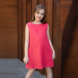 May include: A sleeveless, coral-colored dress. The dress has a simple, straight cut and falls to the mid-thigh. The fabric appears to be linen or a similar lightweight material. The background is a wooden wall.
