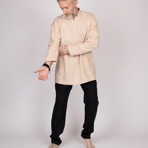 May include: A beige tunic-style shirt with a mandarin collar and long sleeves, paired with black pants. The outfit is simple and elegant, suitable for casual or semi-formal wear. The model is barefoot.