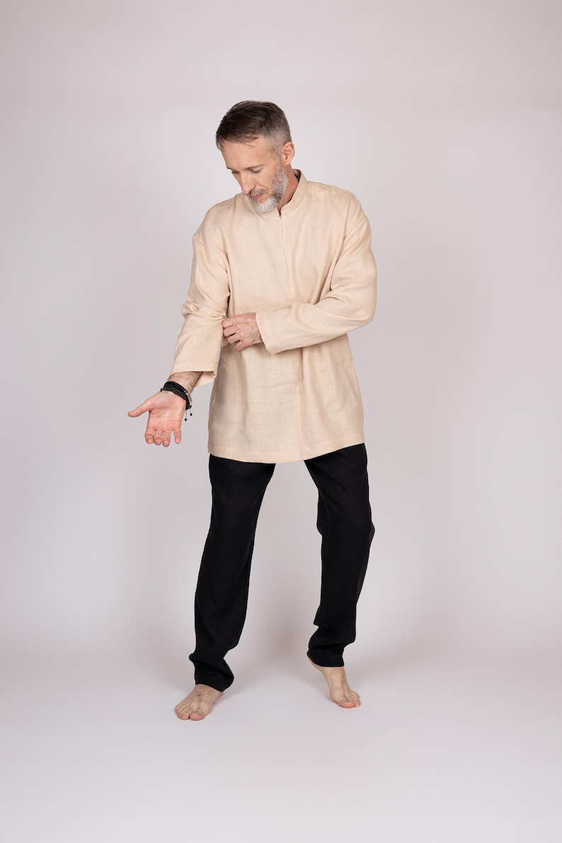 May include: A beige tunic-style shirt with a mandarin collar and long sleeves, paired with black pants. The outfit is simple and elegant, suitable for casual or semi-formal wear. The model is barefoot.