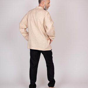 May include: A beige linen shirt with a mandarin collar and long sleeves, paired with black pants. The outfit is simple and classic, suitable for casual wear. The model is standing with their back to the camera.