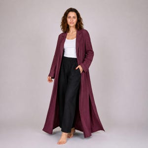 Puede incluir: Un abrigo largo color burdeos con cuello con solapas y aberturas laterales. El abrigo se lleva sobre una camiseta sin mangas blanca y pantalones negros. El atuendo es informal y cómodo.