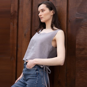 以下が含まれることがあります： スカラップネックラインとウエストにタイディテールが施された、ノースリーブのグレーのリネントップ。青いデニムジーンズと組み合わせています。モデルは木の壁に寄りかかっています。