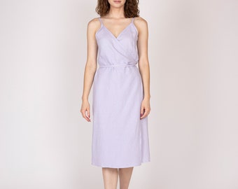 May include: A light purple wrap dress with thin straps and a midi length. The dress has a V-neckline and a matching belt. The model is standing in front of a white background.