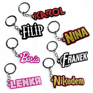 May include: A collection of personalized keychains with names in various colors and fonts. Each keychain has a metal ring and chain. Names include Karol, Filip, Nina, Basia, Franek, Lenka, and Nikodem.