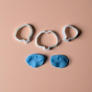 Może przedstawiać: Zestaw białych plastikowych foremek do ciastek w kształcie płatków kwiatów i dwa niebieskie kawałki masy cukrowej w kształcie płatków na jasnoróżowej powierzchni. Foremki mają różne kształty, a kawałki masy cukrowej mają teksturowany wygląd.
