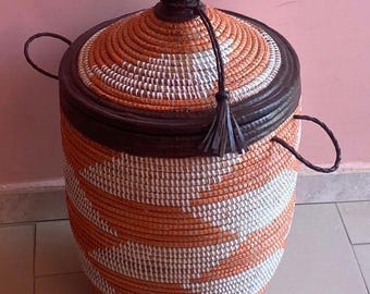 Könnte beinhalten: Ein gewebter Aufbewahrungskorb mit Deckel. Der Korb ist zylindrisch mit braunem Rand und Deckel sowie einem orange-weiß gestreiften Muster. Er hat zwei Griffe und eine Quaste. Der Korb ist etwa 46 cm hoch.