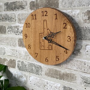 May include: A circular wooden wall clock featuring engraved numerals and a building design. The clock has black hands and is mounted on a brick wall. The clock's diameter is approximately 25 cm.