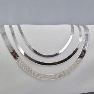 May include: Three silver snake chain necklaces of varying widths, layered on a white surface.