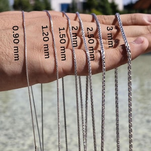 May include: A close-up of a hand holding eight silver chains of different thicknesses. The chains are labeled with their thickness in millimeters: 0.90 mm, 1.20 mm, 1.50 mm, 2 mm, 2.50 mm, 3 mm.