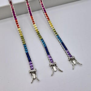 Op de afbeelding: Drie zilveren armbanden met regenboogkleurige vierkante edelstenen. De armbanden zijn in een rij gelegd en de sluitingen zijn zichtbaar.