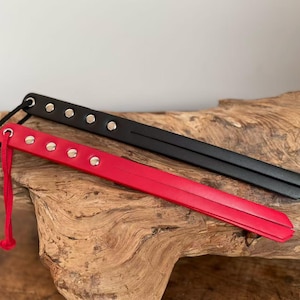 Puede incluir: Dos látigos de cuero con tachuelas plateadas. Un látigo es negro y el otro es rojo. Ambos látigos tienen un lazo en el extremo para sujetar.