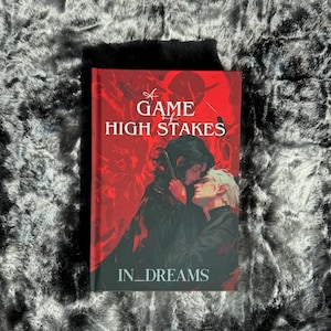 Könnte beinhalten: Ein roter Buchumschlag mit dem Titel "A Game of High Stakes" und dem Untertitel "In_Dreams". Der Umschlag zeigt eine dramatische Illustration von zwei Personen, die sich umarmen.