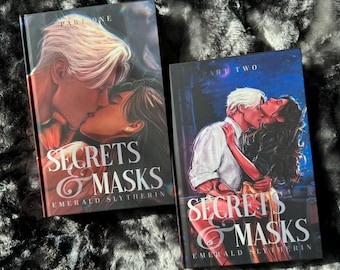 May include: Two books titled "Secrets & Masks" by Emerald Slytherin. The covers show illustrations of a couple kissing. "Part One" is on the first book, and "Part Two" is on the second book. The books are on a dark, textured surface.