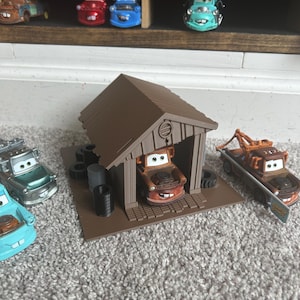 May include: A collection of toy cars, including a brown tow truck and a brown car in a brown garage. The cars are displayed on a shelf and on a gray carpet. The toy cars are from the movie Cars.