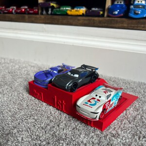 May include: A red plastic podium with three toy race cars. The cars are in first, second, and third place. The first place car is white with blue and red accents and the number 5 on the side. The second place car is black with a purple car behind it. The podium has the words "1st" and "2nd" printed on it.