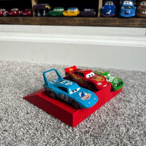 May include: A red, blue, and green toy car racing set with a red ramp. The cars are labeled "95", "43", and "Dinoco". The ramp has the words "2nd" and "1st" printed on it.