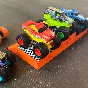 Puede incluir: Una colecci&oacute;n de camiones monstruo de juguete en varios colores, incluyendo rojo, verde y azul, con neum&aacute;ticos grandes. Los camiones se exhiben en un soporte naranja con los n&uacute;meros "ONE" y "TWO" visibles. El soporte tiene un patr&oacute;n de cuadros.