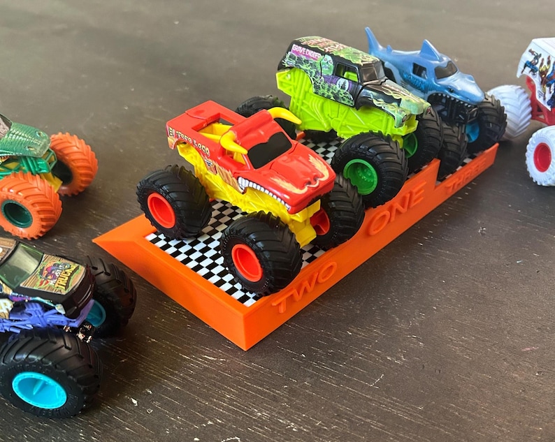 Puede incluir: Una colecci&oacute;n de camiones monstruo de juguete en varios colores, incluyendo rojo, verde y azul, con neum&aacute;ticos grandes. Los camiones se exhiben en un soporte naranja con los n&uacute;meros "ONE" y "TWO" visibles. El soporte tiene un patr&oacute;n de cuadros.