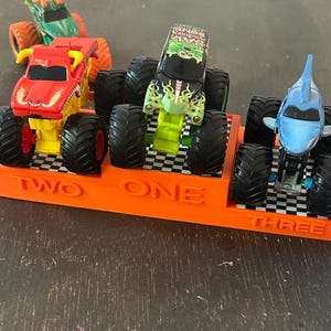 Puede incluir: Un expositor naranja con las palabras "TWO ONE THREE" sostiene tres camiones monstruo de juguete. Los camiones son rojos, verdes y azules, con neum&aacute;ticos negros grandes. El expositor tiene un patr&oacute;n de cuadros.