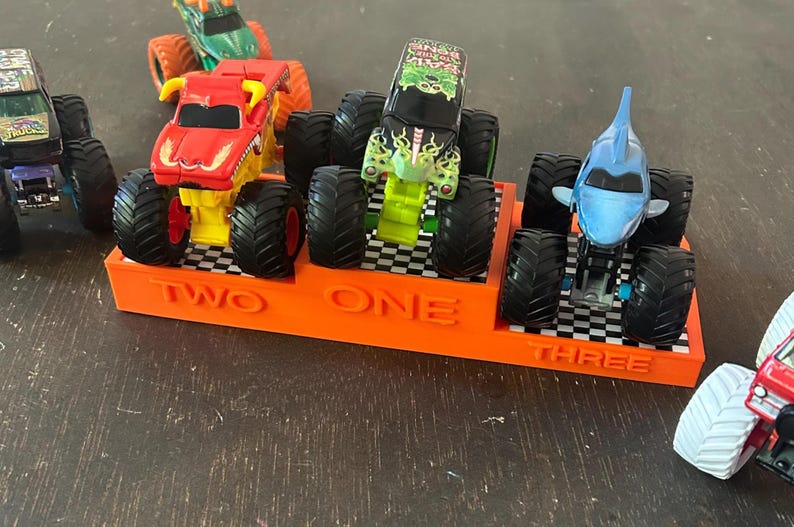 Puede incluir: Un expositor naranja con las palabras "TWO ONE THREE" sostiene tres camiones monstruo de juguete. Los camiones son rojos, verdes y azules, con neum&aacute;ticos negros grandes. El expositor tiene un patr&oacute;n de cuadros.