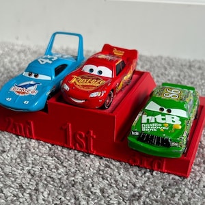 May include: A red, blue, and green toy car racing set with a red podium. The podium has the words "1st", "2nd", and "3rd" printed on it. The cars are from the Disney Pixar Cars movie franchise.