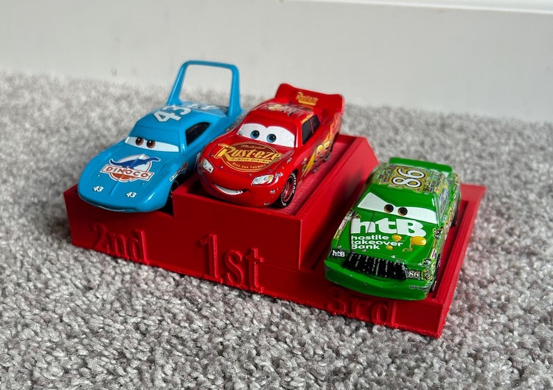 May include: A red, blue, and green toy car racing set with a red podium. The podium has the words "1st", "2nd", and "3rd" printed on it. The cars are from the Disney Pixar Cars movie franchise.