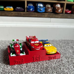 May include: A red race car, a green race car, and a yellow race car on a red podium. The podium has the words "1st", "2nd", and "3rd" printed on it.