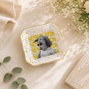 May include: A white ceramic trinket dish with a gold rim, featuring a black and white photo of a child wearing sunglasses and a bandana. The dish is decorated with gold confetti and the words "SENIOR 2026".