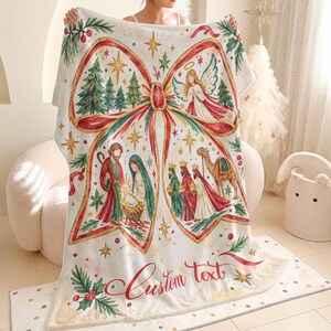 May include: A white fleece blanket featuring a large red ribbon graphic containing a watercolour nativity scene with Mary, Joseph, the baby Jesus, an angel, the three wise men, and a camel. The design includes pine trees, stars, and the text Custom text.