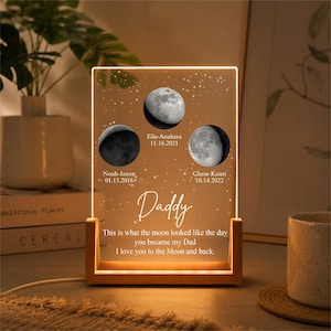 May include: A personalized night light with a wooden base and a clear acrylic panel. The panel features three moon phase images, names, and dates, with the word "Daddy" and a loving message. The light glows with a warm, inviting hue.
