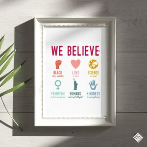 May include: A white framed print with the text "WE BELIEVE" in red. The print features six colorful icons with text below each icon. The icons represent Black Lives Matter, Love is Love, Science is Real, Feminism is for Everyone, Humans are not Illegal, and Kindness is Everything.