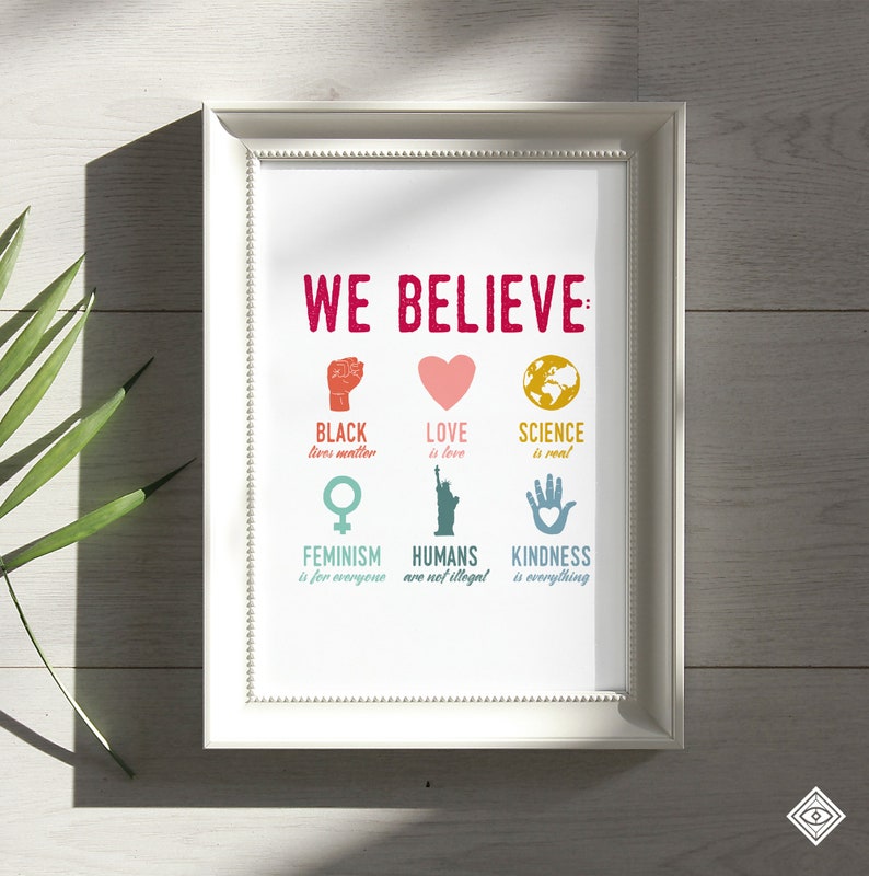 May include: A white framed print with the text "WE BELIEVE" in red. The print features six colorful icons with text below each icon. The icons represent Black Lives Matter, Love is Love, Science is Real, Feminism is for Everyone, Humans are not Illegal, and Kindness is Everything.