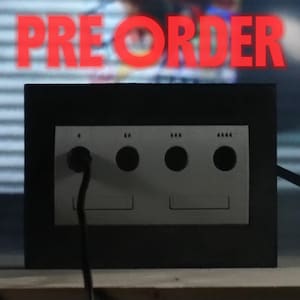 May include: A black video game console front panel featuring four controller ports with circular inputs and two memory card slots below, with the red text PREORDER displayed on the screen behind the console and a black controller to the left.