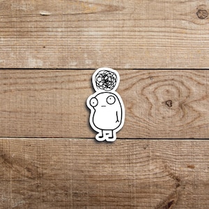 May include: A white sticker depicting a cartoon figure with a bewildered expression. The character has a simple oval body, large eyes, and a scribbled design above its head, representing a thought bubble. The sticker is placed on a wooden surface.