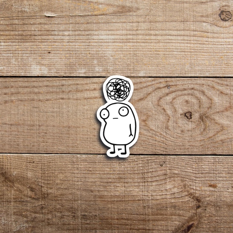 May include: A white sticker featuring a cartoon character with a confused expression. The character has a simple oval body, large eyes, and a scribbled design above its head, suggesting a thought bubble. The sticker is on a wooden surface.