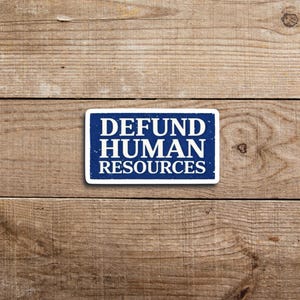 May include: A rectangular sticker with a blue background and white text that reads "DEFUND HUMAN RESOURCES." The sticker has a white border and is placed on a wooden surface.