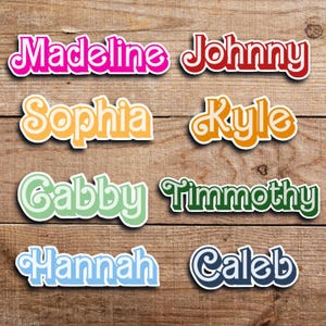 May include: A collection of colorful name stickers on a wooden surface. Names include Madeline, Johnny, Sophia, Kyle, Gabby, Timothy, Hannah, and Caleb. Each name is in a different color and font, with a white border.
