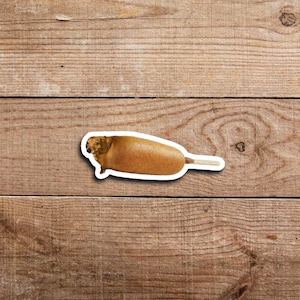 May include: A sticker depicting a dachshund dog designed to resemble a corn dog. The dog is brown with a white border, and the corn dog batter is golden brown. The stick is white.